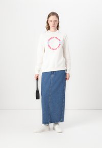 Kremet sweatshirt med rød og blå tekst "TOMMY JEANS NEW YORK CITY", satt sammen med et langt blått denimskjørt og hvite sneakers, holder en svart veske.