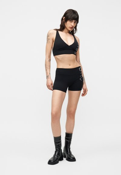 Femme tatouée portant un brassière de sport noire, un short taille haute noir avec texte blanc, et des bottes noires mi-mollet, debout devant un fond blanc.