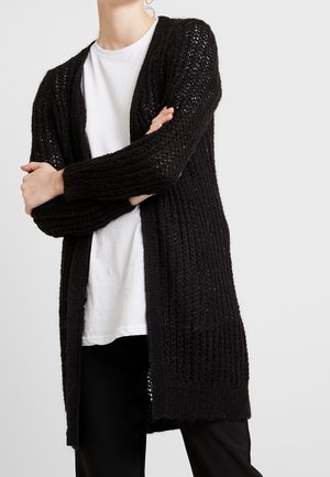 Personne portant un long cardigan noir en maille sur un T-shirt blanc et un pantalon noir, les bras croisés devant un fond uni.
