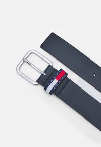 Cinturón de cuero azul marino con hebilla rectangular plateada, que presenta una etiqueta de tela en rojo, blanco y azul con el texto "TOMMY JEANS". Textura suave.