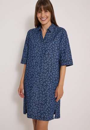 Femme souriante portant une robe bleue mi-longue avec un imprimé léopard discret et des manches trois-quarts, debout devant un fond clair uni.