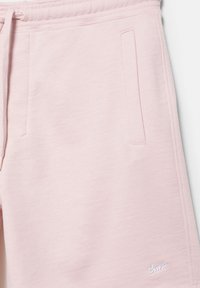 Gros plan sur un pantalon de survêtement rose clair montrant la poche latérale avant, la ceinture à cordon de serrage et un petit logo blanc cousu près de l'ourlet.