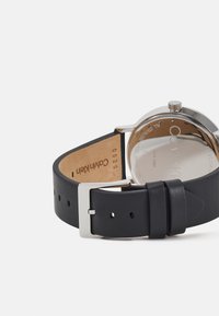 Montre à bracelet en cuir noir avec un boîtier en acier inoxydable. Présente une texture en cuir lisse, une boucle en métal et un gravure intérieure de marque.