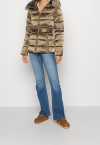 Donna che indossa un giubbotto imbottito bronzo lucido con cappuccio in pelliccia e cintura, jeans a campana azzurri chiari e scarpe mocassino marroni.