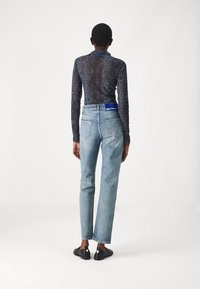 Langsleeve top met een donker slangenhuidpatroon, gecombineerd met lichtblauwe high-waisted tapered jeans en zwarte instappers met gouden accenten.