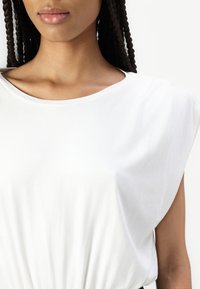 Haut en coton blanc avec un col rond et des manches courtes structurées ; la texture lisse et la coupe ample à la taille créent une silhouette décontractée.