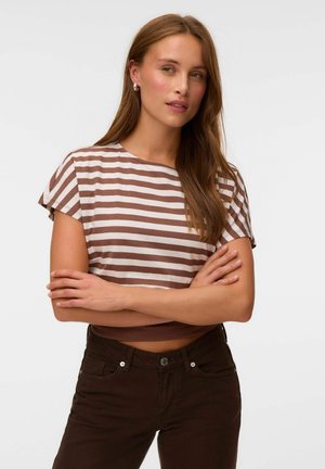 Femme aux longs cheveux bruns portant un t-shirt rayé marron et blanc à manches courtes et un pantalon marron foncé, les bras croisés.
