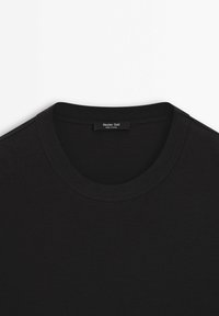 T-shirt noir à col rond avec une étiquette visible cousue à l'intérieur du col portant la mention "Massimo Dutti Made in China" sur fond blanc.