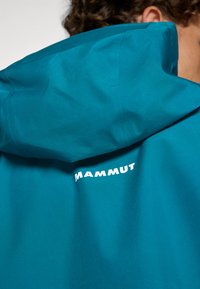 Teal waterproof jacket with a hood, featuring a visible logo in white on the back. Smooth texture, no patterns or additional hardware.