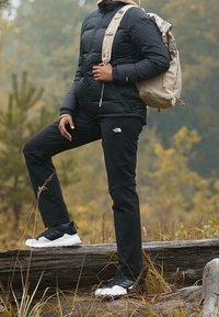 Puffjacka i svart, svarta byxor med logotyp, beige ryggsäck och flerfärgade sneakers; placerad i en naturlig utomhusmiljö med träd.