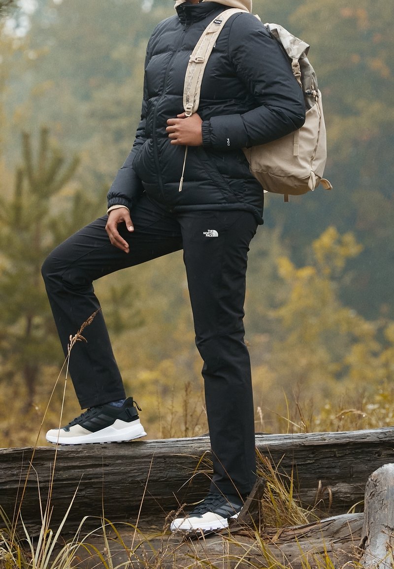 Puffjacka i svart, svarta byxor med logotyp, beige ryggsäck och flerfärgade sneakers; placerad i en naturlig utomhusmiljö med träd.