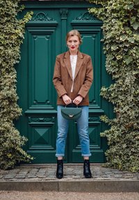 Brauner Wildlederblazer über einem weißen Oberteil mit Schnürdetails, hellblaue hochgekrempelte Jeans, schwarze Ankle Boots und eine grüne Handtasche. Hintergrund mit grüner Tür.