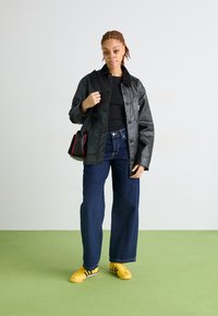 Giacca di pelle nera, top nero aderente, jeans blu larghi, sneaker gialle con strisce, che tiene una borsa nera con accenti rossi, in piedi su un pavimento verde.