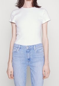 Žena v jednoduchém bílém tričku s krátkým rukávem zasunutém do světle modrých džínsových kalhot s vysokým pasem na bílém pozadí.