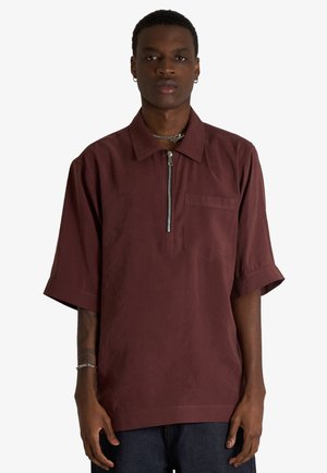 Uomo che indossa una camicia bordeaux a maniche corte larga con zip frontale e taschino sul petto, abbinata a pantaloni scuri, in piedi su sfondo bianco.