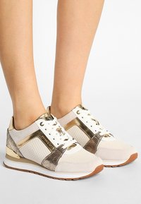 Jasnobeżowe sneakersy z metaliczno-złotymi akcentami, teksturowanymi bocznymi panelami i białymi sznurówkami. Gumowa podeszwa, gładkie i błyszczące wykończenie, elegancki design.