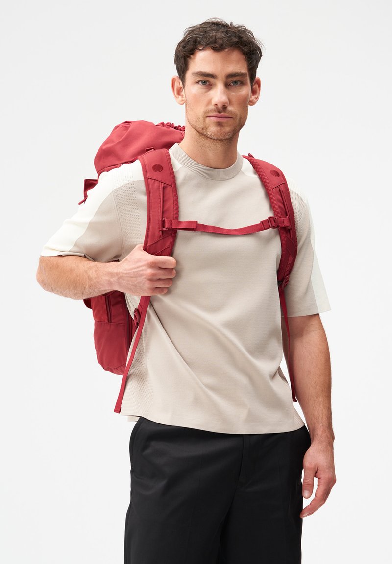 Homme aux cheveux courts et bouclés portant une chemise beige à manches courtes et un pantalon noir, avec un grand sac à dos rouge équipé d'une sangle pectorale.