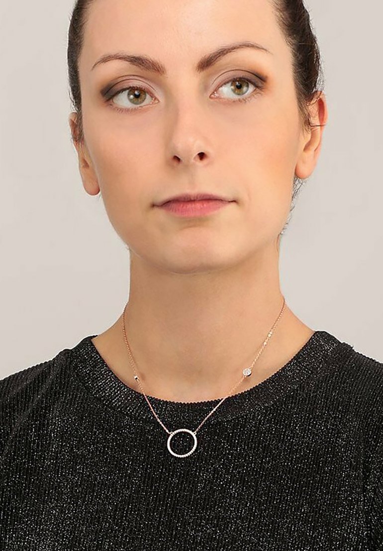 Collana in oro rosa con un pendente circolare, decorato con piccoli accenti, indossata su un top scuro e texturizzato. Design della catena e del gancio semplice.