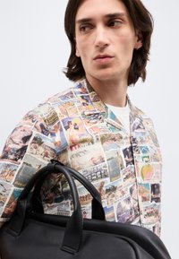 Νεαρός με μακριά μαλλιά μέχρι τους ώμους, φορώντας πολύχρωμο πουκάμισο με μοτίβο καρτ ποστάλ, κρατώντας μαύρη δερμάτινη τσάντα, κοιτάζει προς το πλάι.