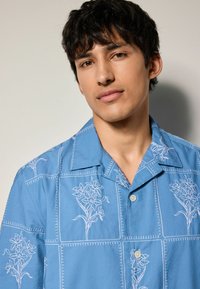 Blauwe short-sleeve shirt met witte bloemenborduursels, vierkante patronen en een knoopsluiting aan de voorkant. De stof lijkt licht en soepel te zijn.