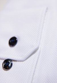 Witte textiele stof met een close-up van een gevouwen shirtkraag met twee zwarte knopen en zichtbare stiksels.