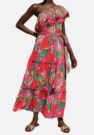 Nainen pukeutuneena vaaleanpunaiseen olkaimettomaan maksimekkoon, jossa on trooppinen kukkakuvio ja tupsureunus, yhdistettyinä kultaisiin litteisiin sandaaleihin.