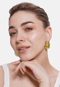 Orecchini geometrici dorati con un design testurizzato e scultoreo. La modella si tocca il mento, indossando una semplice maglietta bianca e un trucco naturale.