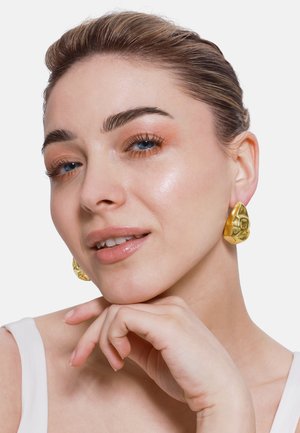 Goldene geometrische Ohrringe mit einem strukturierten, skulpturalen Design. Die Model berührt ihr Kinn und trägt ein einfaches weißes Oberteil sowie ein natürliches Makeup.