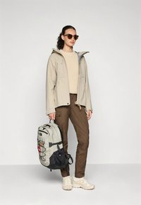 Beige vattentät jacka, ljus tröja, bruna byxor och beiga vandringsskor. Grå och svart ryggsäck med remmar, unika designdetaljer.