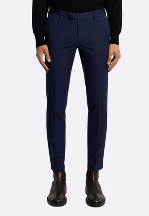 Uomo che indossa pantaloni slim-fit blu navy, maglia nera e stivaletti marrone scuro, in piedi con le mani rilassate ai lati.