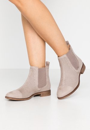 Beine überschlagen mit beigen Wildleder-Stiefeletten mit perforiertem Design, seitlichen elastischen Einsätzen und niedrigen Holzabsätzen auf weißem Boden.