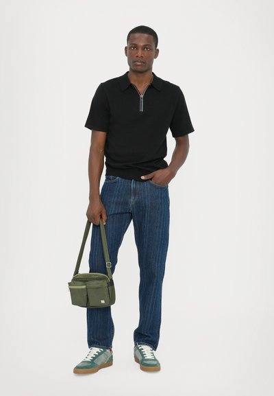 Polo negru cu mâneci scurte și fermoar, blugi cu model albastru, adidași verzi și o geantă de umăr verde cu un buzunar frontal.