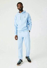 Uomo che indossa un hoodie azzurro chiaro e pantaloni jogger abbinati, con sneaker bianche e nere, in piedi contro uno sfondo bianco uniforme.