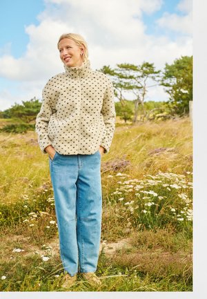 Jeune femme portant une veste polaire légère à motifs et un jean bleu se tient debout en souriant, les mains dans les poches, dans un champ herbeux avec des fleurs sauvages.