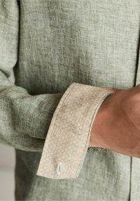 Osoba regulująca beżowy, wzorzysty mankiet na zielonej, teksturowanej koszuli z długim rękawem, pokazująca szczegóły tkaniny i szwy.