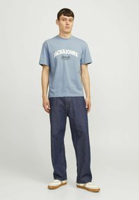 Camiseta de algodón de color azul claro con el logotipo "JACK & JONES" impreso en el frente, combinada con jeans de mezclilla azul oscuro y zapatillas blancas.