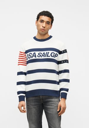 Άνδρας που φοράει λευκό και ναυτικό μπλε ριγέ πουλόβερ με το κείμενο «USA SAIL GP» και κόκκινες ρίγες στο μανίκι, συνδυασμένο με σκούρο γκρι τζιν, στέκεται μπροστά σε λευκό φόντο.