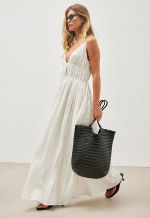 Woman in flowing white dress and black sandals carries large woven black tote bag, wearing dark sunglasses, walking against plain background.