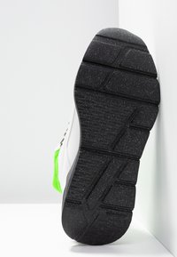 Semelle en caoutchouc noire texturée d'une chaussure de sport blanche et verte positionnée verticalement contre un mur d'angle blanc.