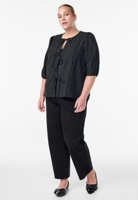 Ženska velikih konfekcijskih številk, oblečena v črno bluzo s črtami in zavezovanjem spredaj, črne hlače naravnega kroja ter črne lakirane loaferje, stoji pred belo ozadjem.