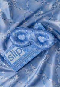 Maschera per dormire in seta blu con motivo trapuntato e design a fiocco, abbinata a una scatola coordinata con il nome del marchio "slip." Texture satinata.