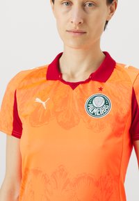 Obleka polo kratkih rukavov v oranžni in rdeči barvi, z teksturiranim cvetličnim vzorcem, Puma logotipom na desnem ramu in emblemom Palmeiras na prsih.