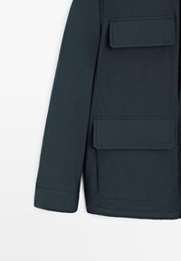 Donkerblauwe buitenjas met een gladde textuur, lange mouwen en twee voorzakken met klep en stikdetails.