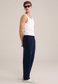 Jūras zila plīša bikses, apvienotas ar baltu bezpiedurkņu blūzi un baltām sporta kedām. Modelis stāv sānis, parādot atbrīvoto piegriezumu un auduma tekstūru.