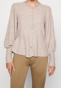 Cárdigan de punto color beige claro con mangas largas abullonadas y dobladillo acampanado, combinado con pantalones de talle alto de color marrón claro con pliegues.