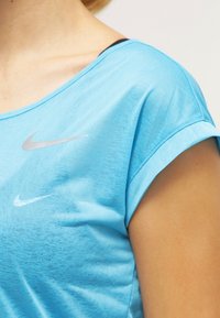 Světle modrý atletický krátký rukáv Nike s hladkou texturou, kulatým výstřihem a dvěma stříbrnými logy Nike na hrudi. Pod ním je viditelná černá vrstva.