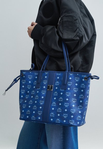 Osoba v čiernej bunde a džínsoch drží veľkú modrú tašku MCM s opakujúcimi sa logami a koženými rúčkami.