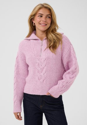 Jeune femme souriante portant un pull torsadé violet clair avec un col à demi-fermeture éclair et un pantalon foncé sur un fond uni.