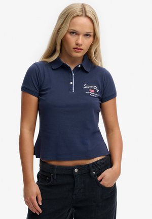 Lyshåret kvinde iført navyblå cropped polo med SuperDry-logo og sorte fløjlsbukser, den ene hånd i lommen, neutralt ansigtsudtryk.
