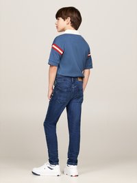 Polo blu con strisce rosse e bianche sulle maniche, abbinato a jeans blu scuro e sneakers bianche. Focalizzato su un look casual e fit rilassato.